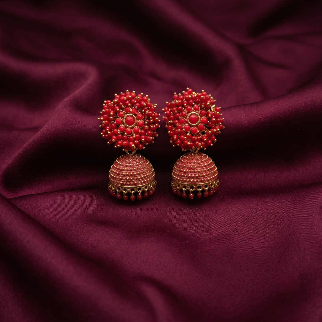 A pair of traditional Indian jhumka earrings with red beaded floral tops and textured red bell-shaped drops, displayed on a draped dark burgundy fabric background.