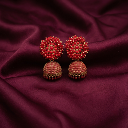 A pair of traditional Indian jhumka earrings with red beaded floral tops and textured red bell-shaped drops, displayed on a draped dark burgundy fabric background.