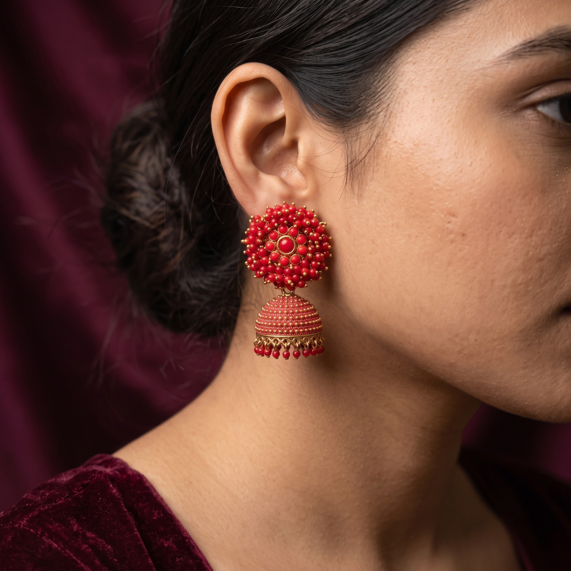 A close-up of a woman's ear wearing a red and gold beaded jhumka earring against a dark burgundy background.