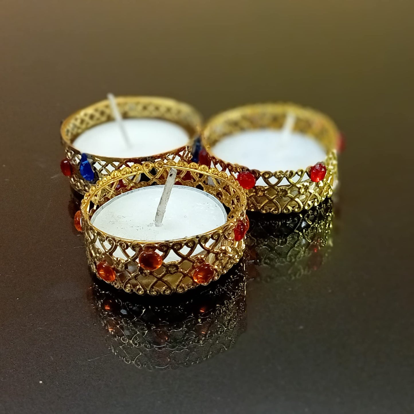 Three small candles in decorative gold holders with red gemstones on a reflective surface.