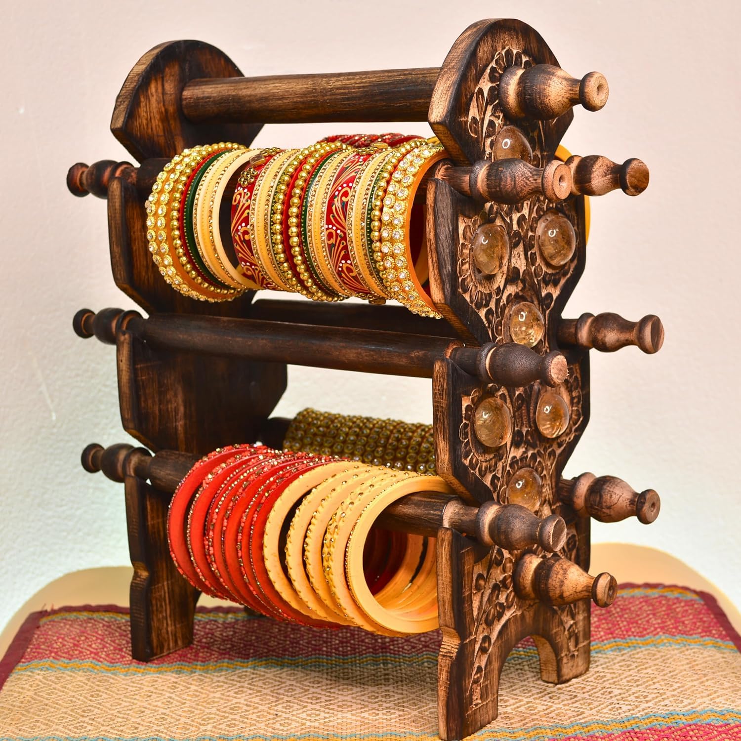 A dark carved wooden bangle rack holding multiple rows of ornate gold, red, and yellow bangles, photographed against a plain light background with a woven mat underneath.