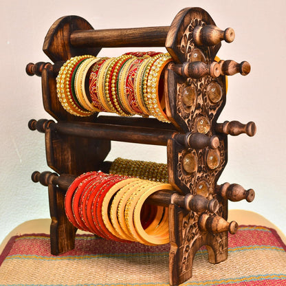 A dark carved wooden bangle rack holding multiple rows of ornate gold, red, and yellow bangles, photographed against a plain light background with a woven mat underneath.