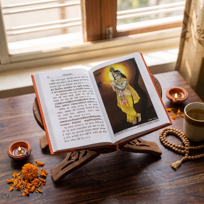 An open book on a wooden stand on a dark wooden table, with Devanagari text on both pages and a printed illustration of a child in a yellow garment on the right page. The scene is styled with lit oil lamps, orange flower petals, a cup of tea, and a beaded necklace near a window with blinds.