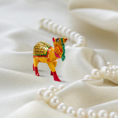 A small hand-painted camel figurine in yellow, red, green, and gold, styled on draped white satin fabric with pearl beads in the foreground and background.