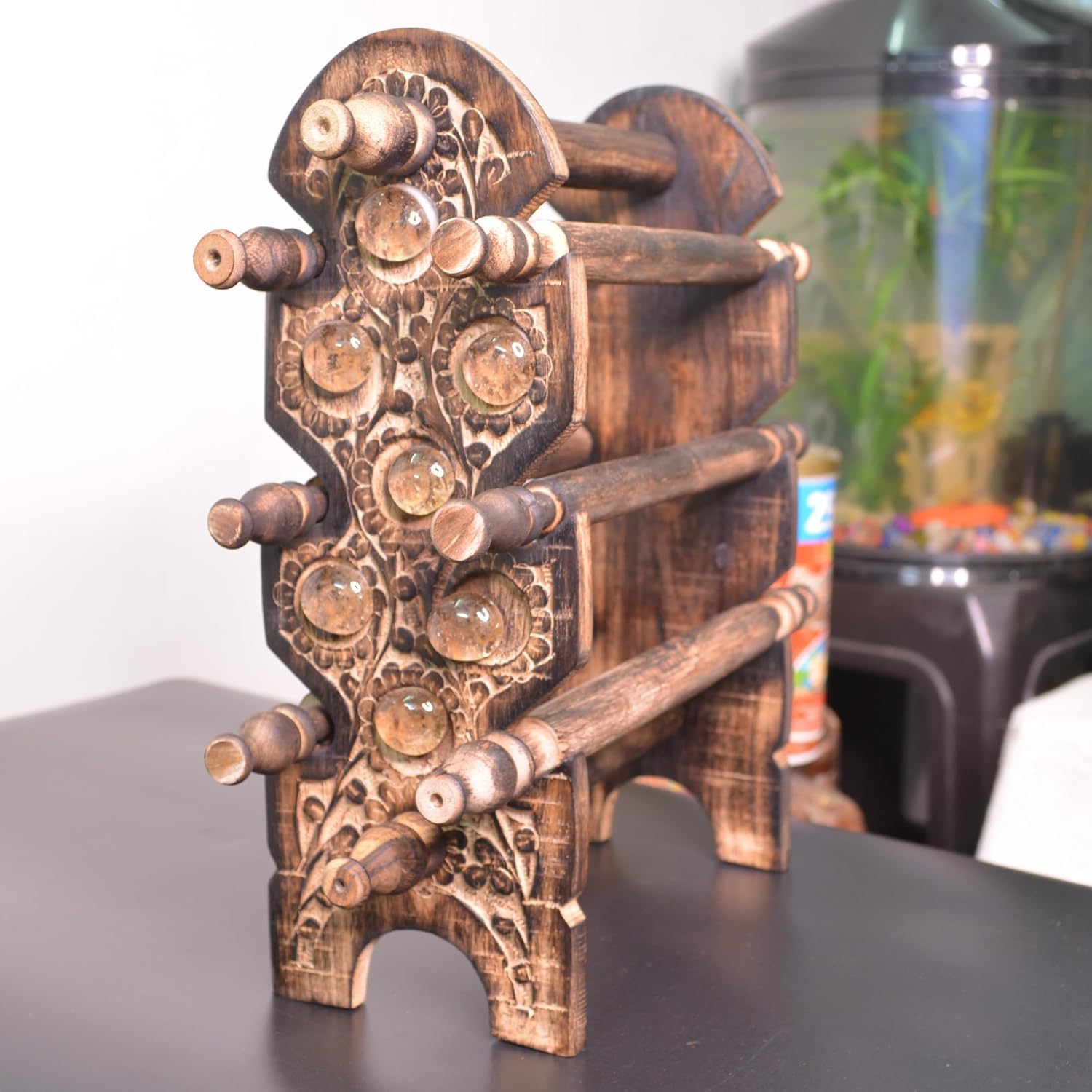 A dark brown carved wooden decorative stand with multiple horizontal rods and circular carved details, photographed on a dark tabletop with a blurred indoor background including a fish tank.