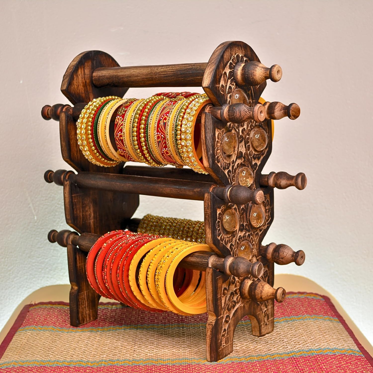 An ornate dark brown carved wooden bangle rack holding multiple rows of colorful bangles, including yellow, red, and gold-toned styles, against a plain light background.