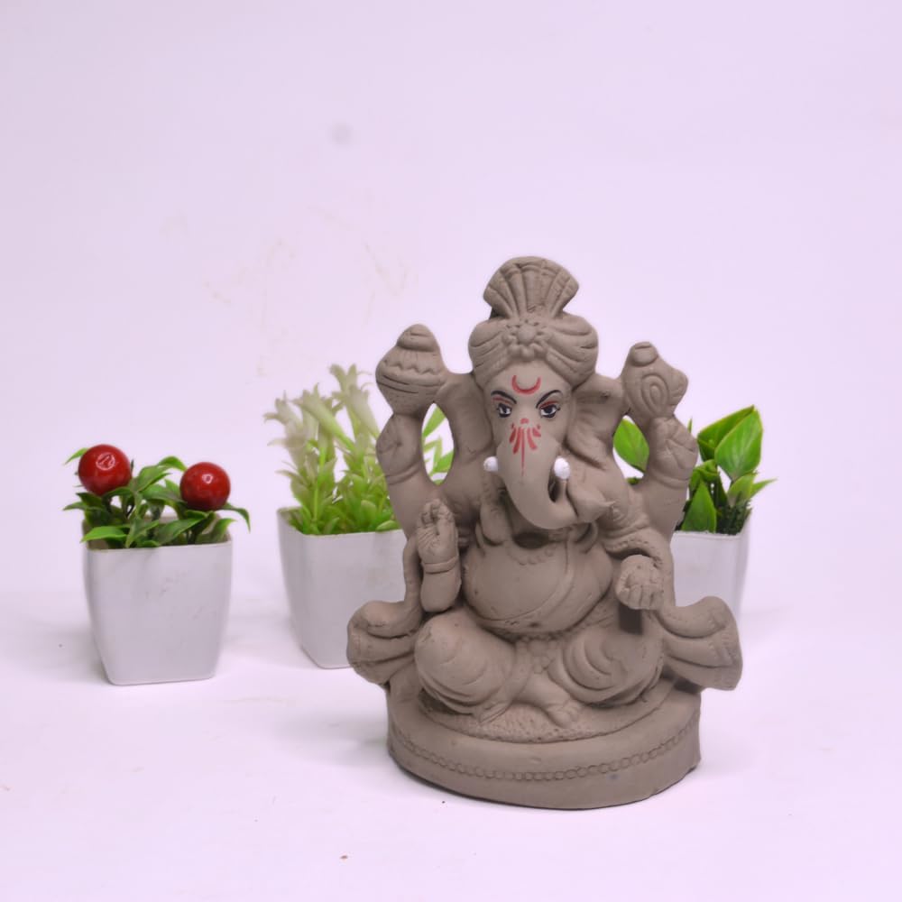A gray clay Ganesha statue seated on a circular base, photographed against a plain light background with three small potted plants behind it.