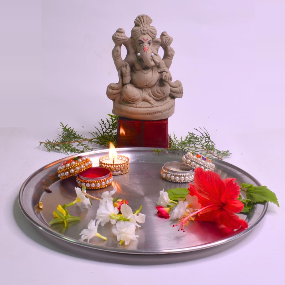 A clay Ganesha statue displayed on a red pedestal behind a round silver tray adorned with lit diya candles, flowers, and small decorative items, set against a plain light background.