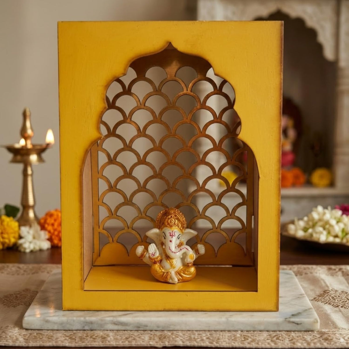 A decorative yellow wooden Ganesha display box with an arched cutout design, shown on a marble board with a brass diya and flowers in a styled indoor setting.