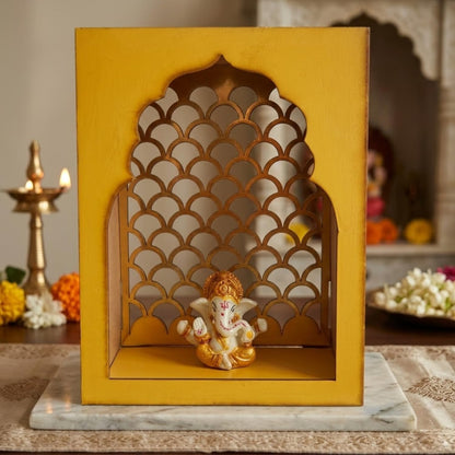 A decorative yellow wooden Ganesha display box with an arched cutout design, shown on a marble board with a brass diya and flowers in a styled indoor setting.
