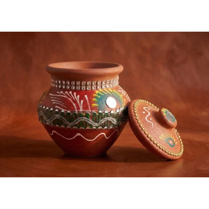 A terracotta clay pot with a matching lid, decorated with hand-painted patterns in green, yellow, red, and white, and accented with small silver mirrors, set against a warm brown background.