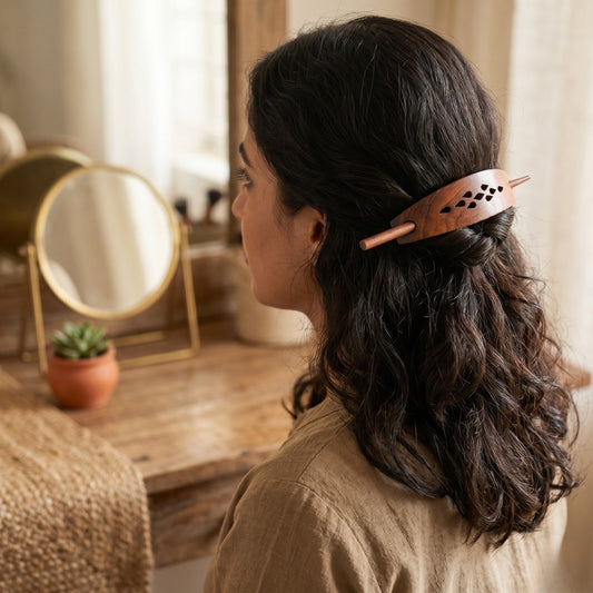 A woman with dark curly hair is shown from the side profile, wearing a copper-toned wooden hair claw clip with decorative cutouts. She is wearing a beige linen top in a warm, softly lit room with a wooden vanity table and a small mirror in the background.