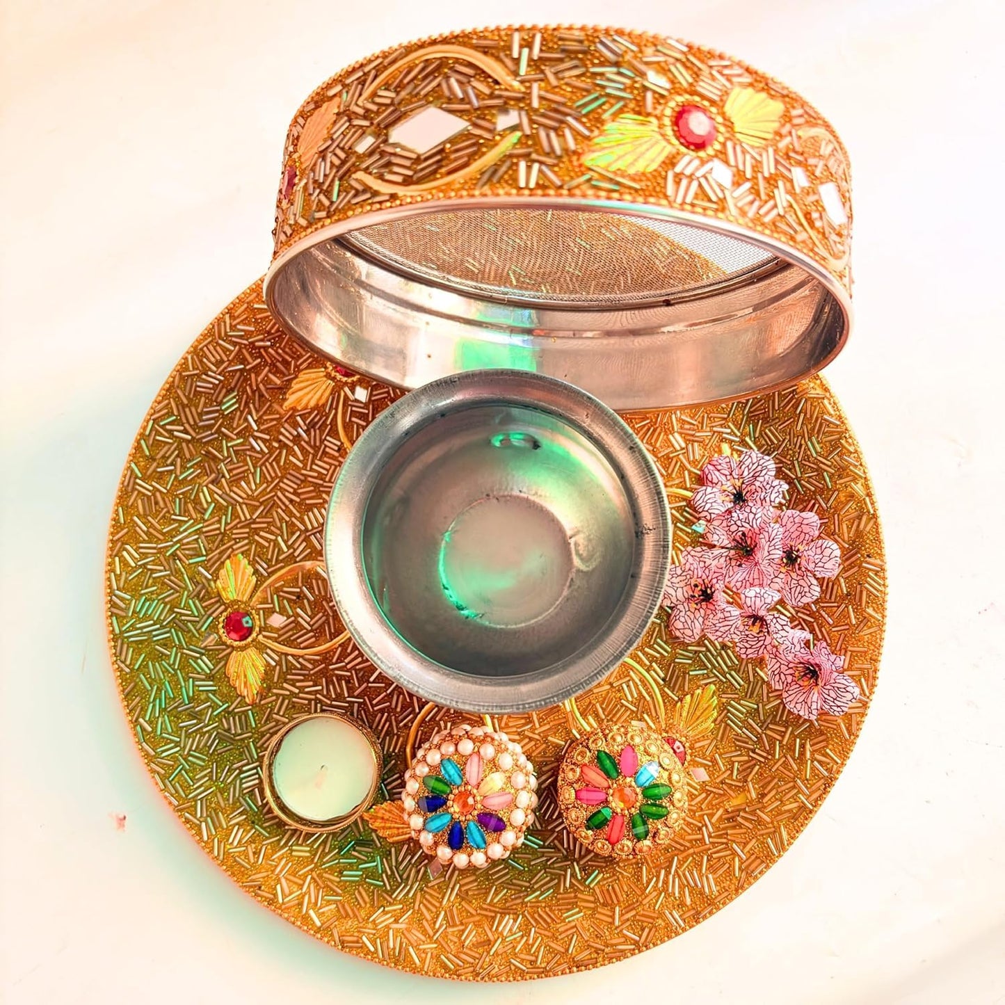 An ornate gold-toned metal diya lamp holder set with a matching tray, open lid, small bowl, decorative floral embellishments, and two lit candles on a plain light background.