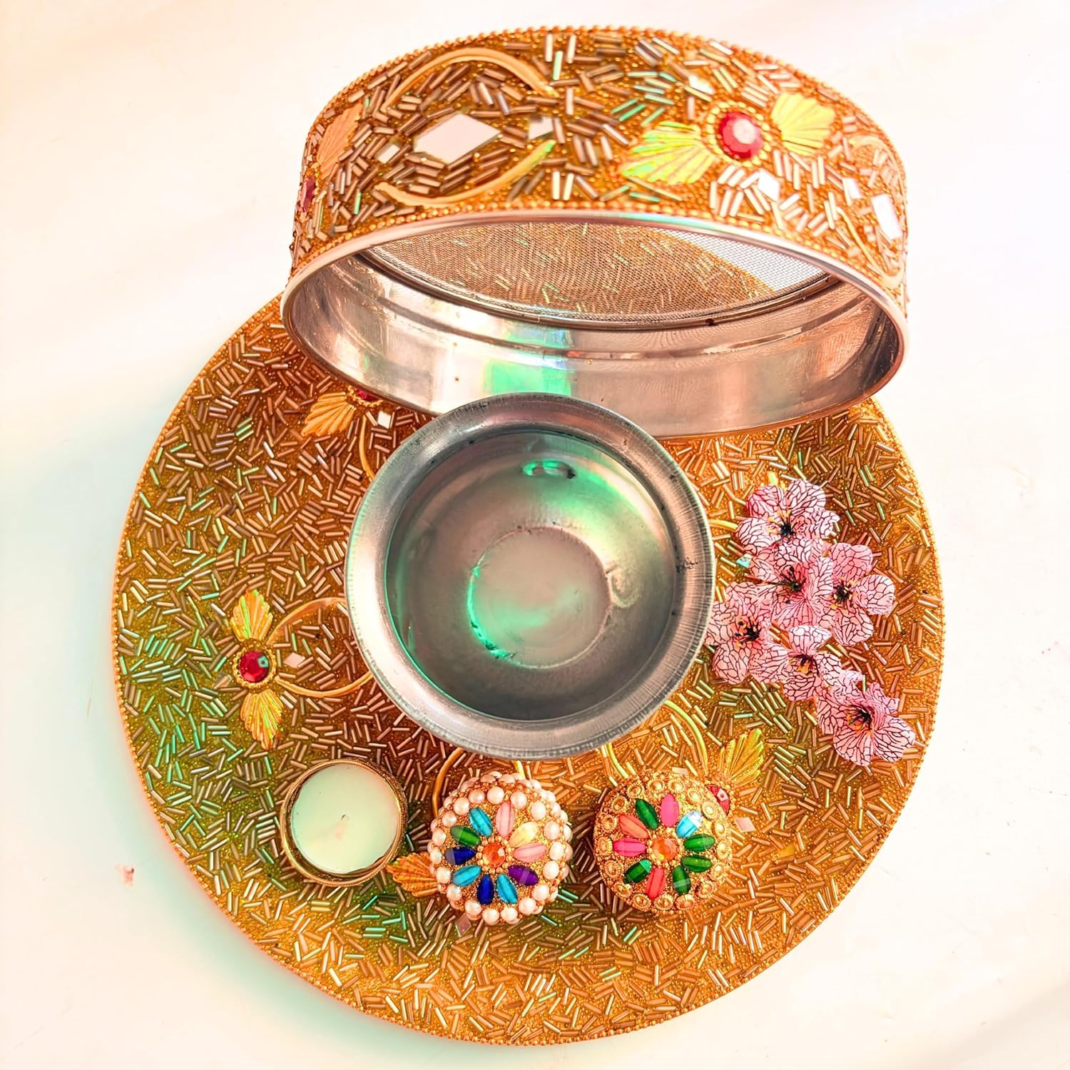 An ornate gold-toned metal diya lamp holder set with a matching tray, open lid, small bowl, decorative floral embellishments, and two lit candles on a plain light background.
