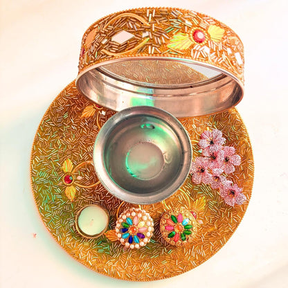 An ornate gold-toned metal diya lamp holder set with a matching tray, open lid, small bowl, decorative floral embellishments, and two lit candles on a plain light background.