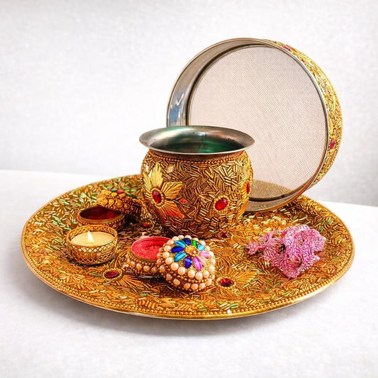 An ornate gold-toned traditional ritual tray set on a white surface, featuring a decorated pot with a lid, a lit tealight candle, a small bowl, a colorful round ornament, and pink flowers.