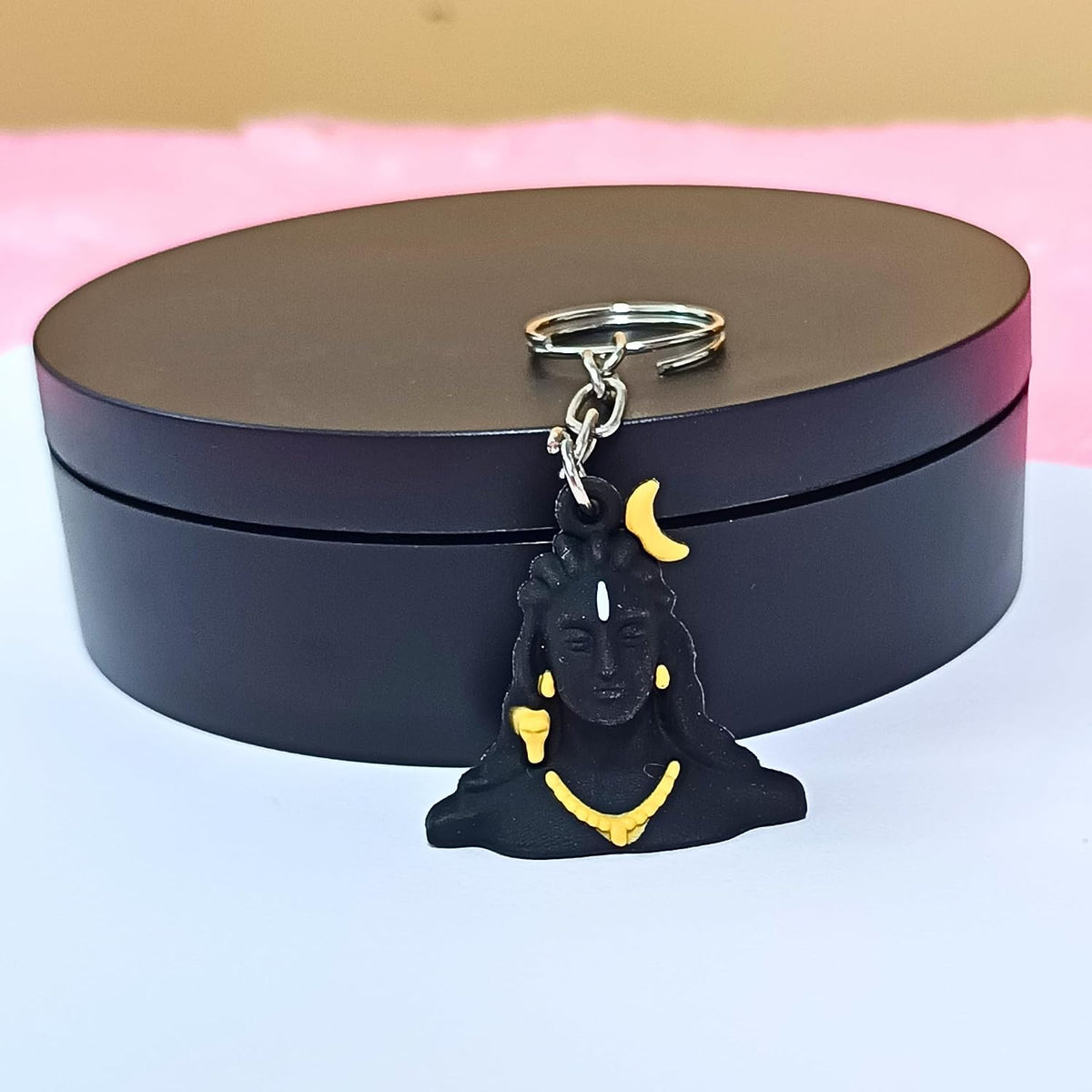 A black Shiva keychain with yellow crescent moon and necklace details, resting on a round black gift box against a pink and white background.