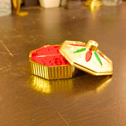 A small gold-toned decorative box with a lid, filled with bright red powder, placed on a dark wooden surface.