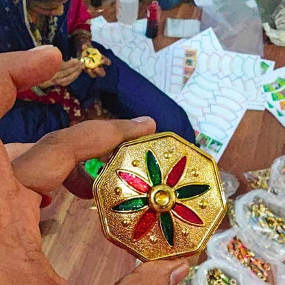 A hand holds a small octagonal gold-toned decorative piece with a flower motif in red and green enamel, while another person in blue clothing works in the background among papers and jewelry components.