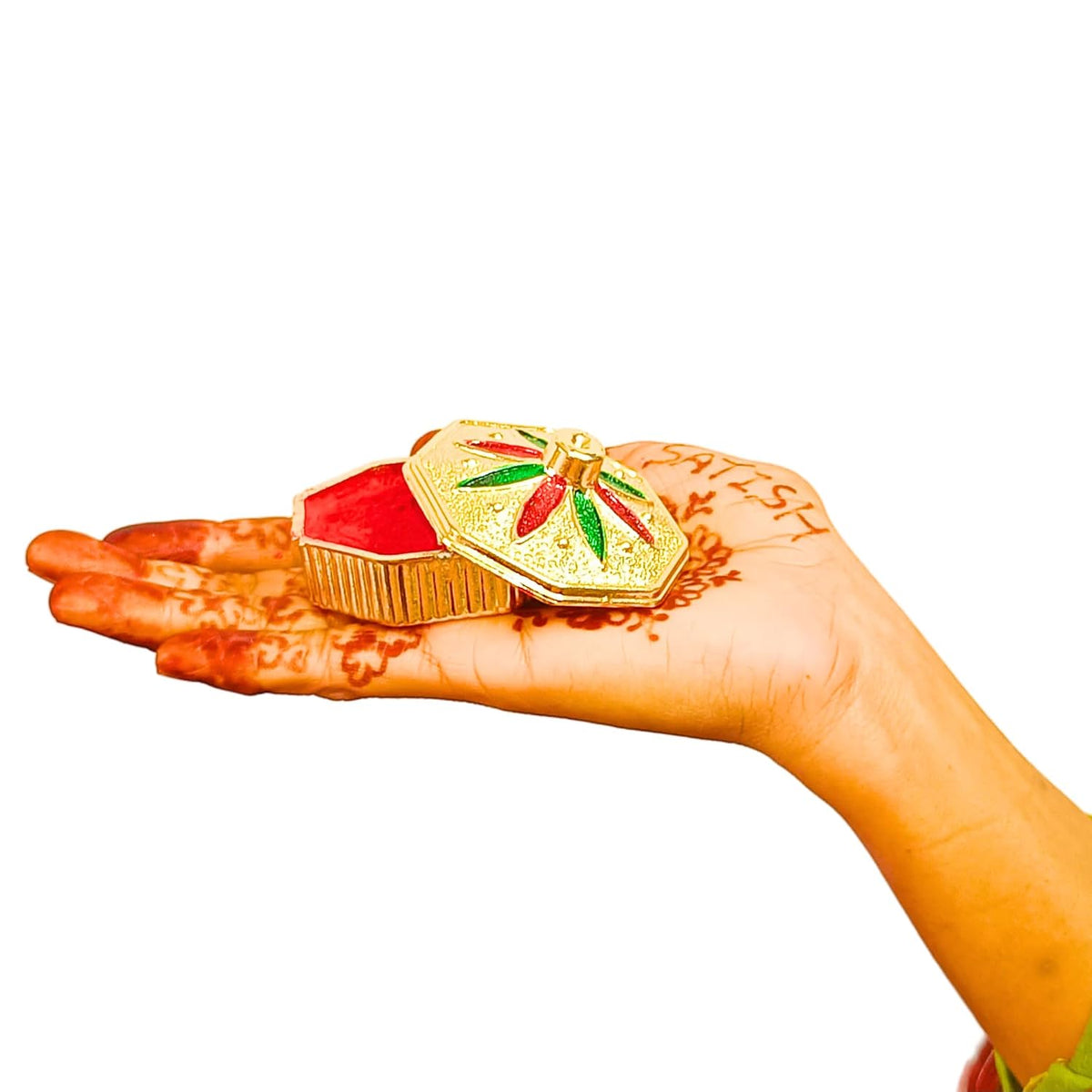 A hand with henna painted the word 'MEST' holds a small ornate gold box with a red interior and a decorative lid featuring red and green floral details, against a plain white background.