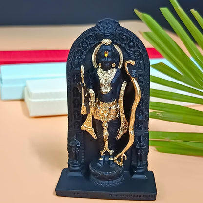 A black and gold Lord Hanuman statue standing in front of an arched black frame, displayed on a beige surface with blurred boxes and green leaves in the background.