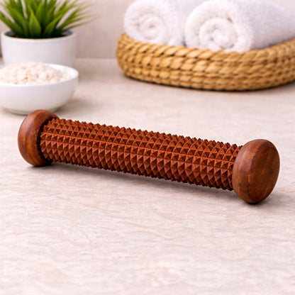 A wooden massage roller with a textured, diamond-patterned surface and rounded ends, resting on a light beige stone surface with spa props in the background.