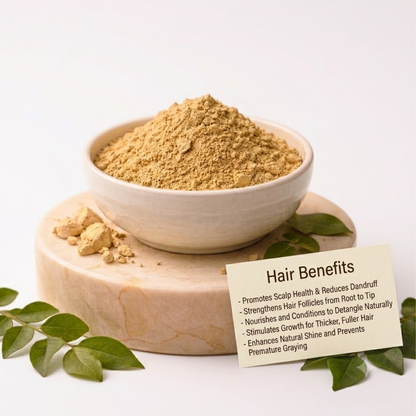 A small bowl filled with light brown powder sits on a round wooden stand, with scattered green leaves around it and a small card showing hair benefits text in front. The setup is photographed on a clean white background.