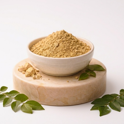 A small white bowl filled with light tan powder sits on a round beige stone slab, with scattered loose pieces and green leaves around it on a clean white background.