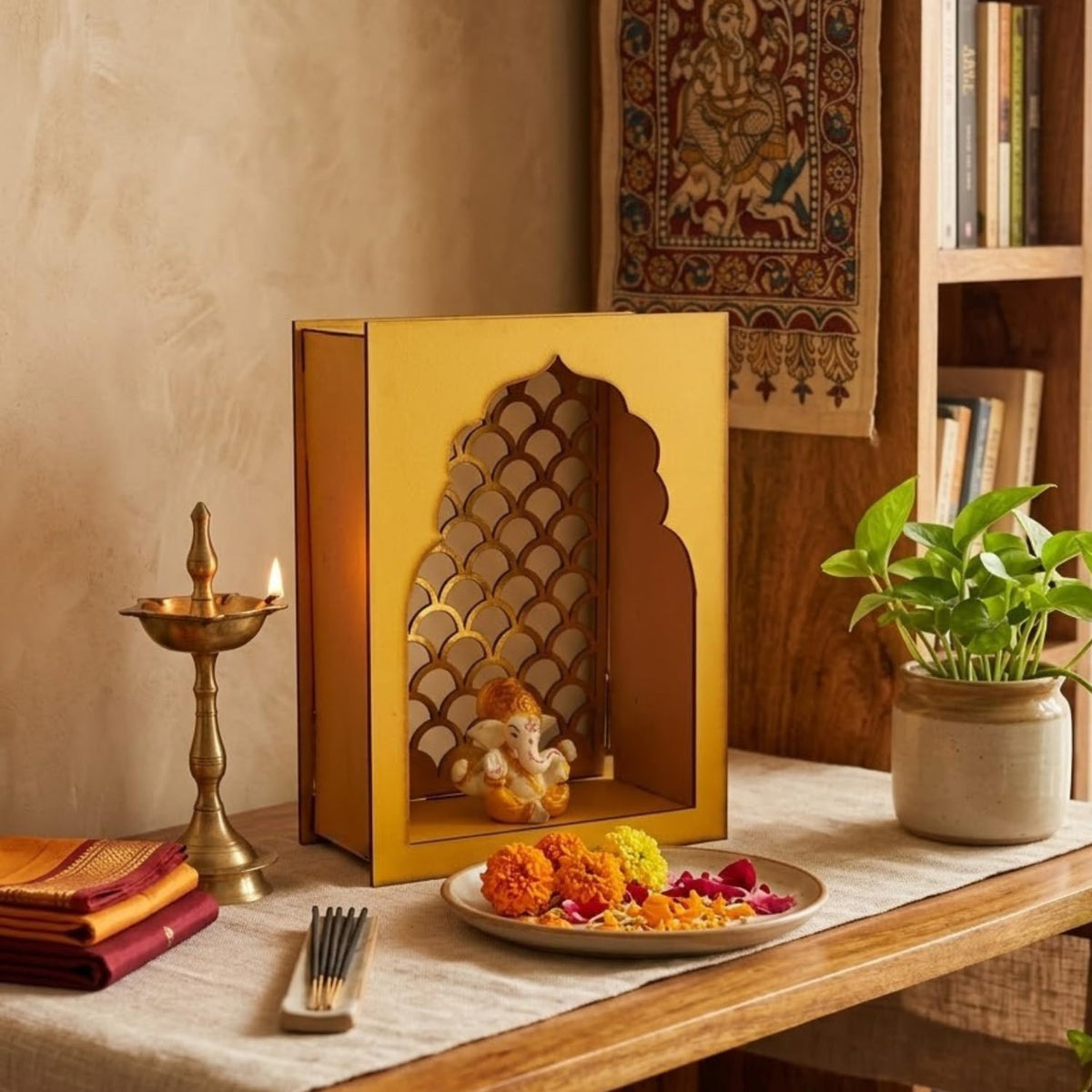 A golden arched decorative box with a cutout pattern and a small Ganesha figurine inside, displayed on a wooden table alongside a brass lamp, folded cloth, incense, and a potted plant in a warm indoor setting.