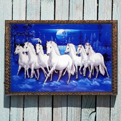A framed wall art piece showing a group of white horses running through a snowy night landscape under a full moon, mounted on a light weathered wood plank wall.