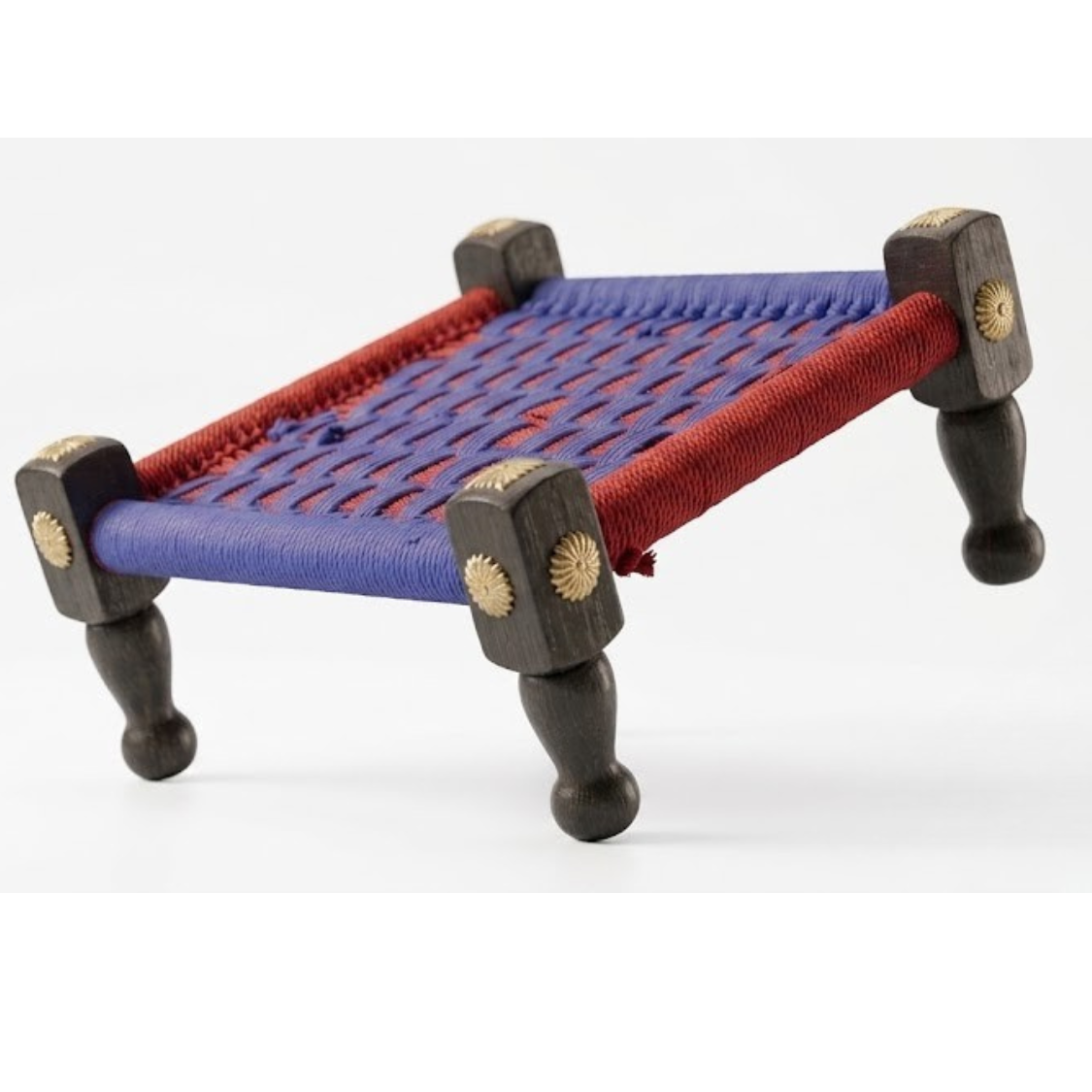 A miniature wooden charpai-style bed or platform with a blue and red woven rope seat, dark brown turned legs, and brass star accents on the corner posts, shown on a white background.
