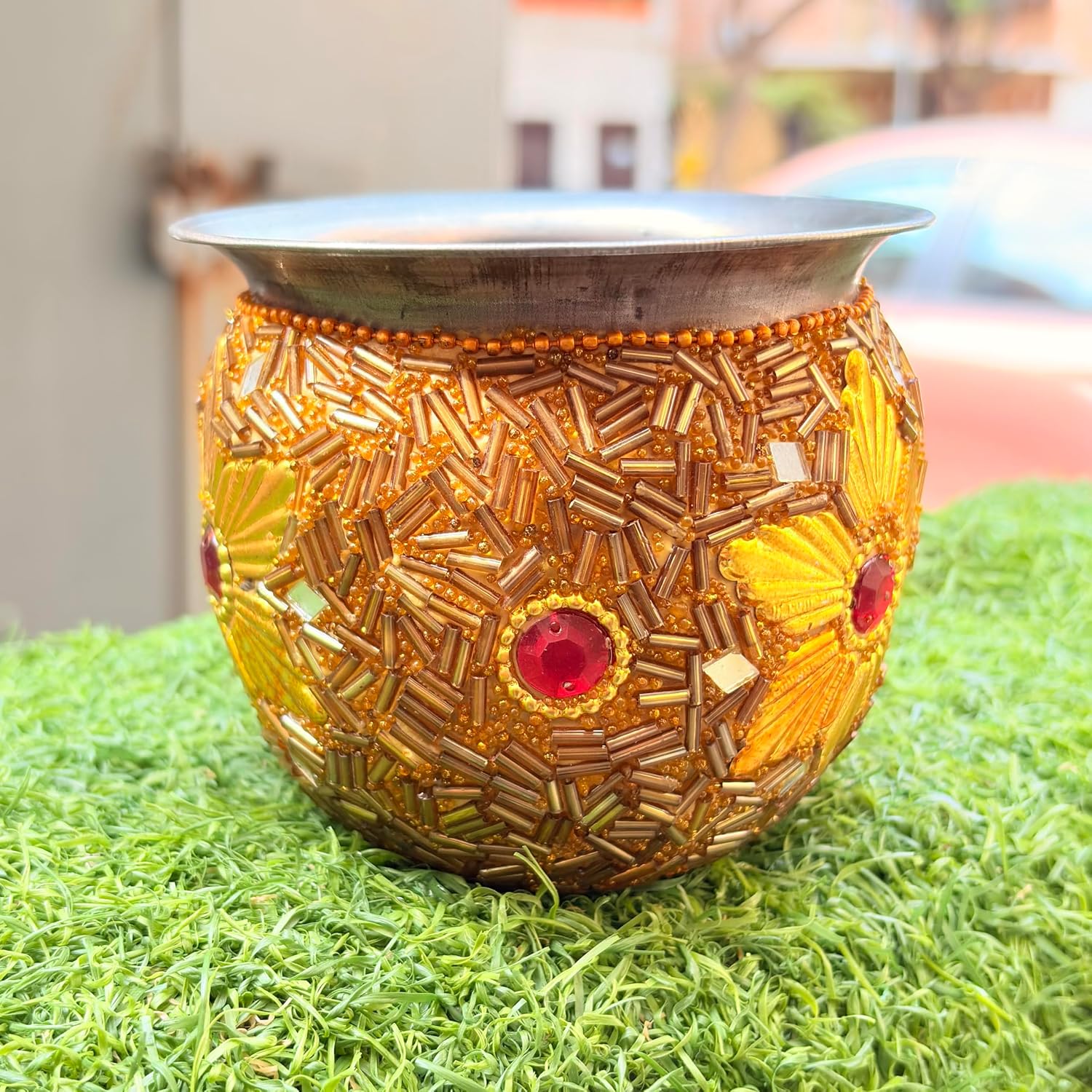 A decorative brass oil lamp with intricate gold and yellow floral beadwork and red gemstone accents, placed on artificial green grass with a blurred outdoor background.