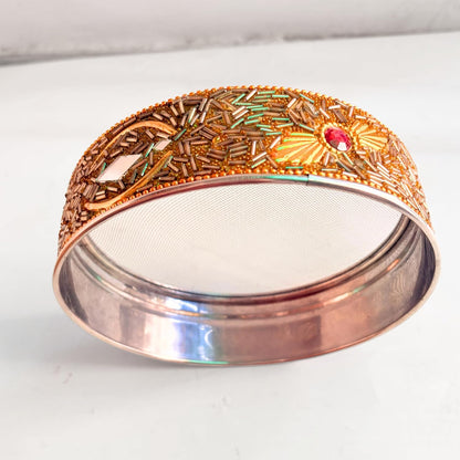 A copper-colored metal bangle bracelet with intricate gold-toned filigree, green and yellow enamel details, and a small red gemstone, shown on a light gray background.