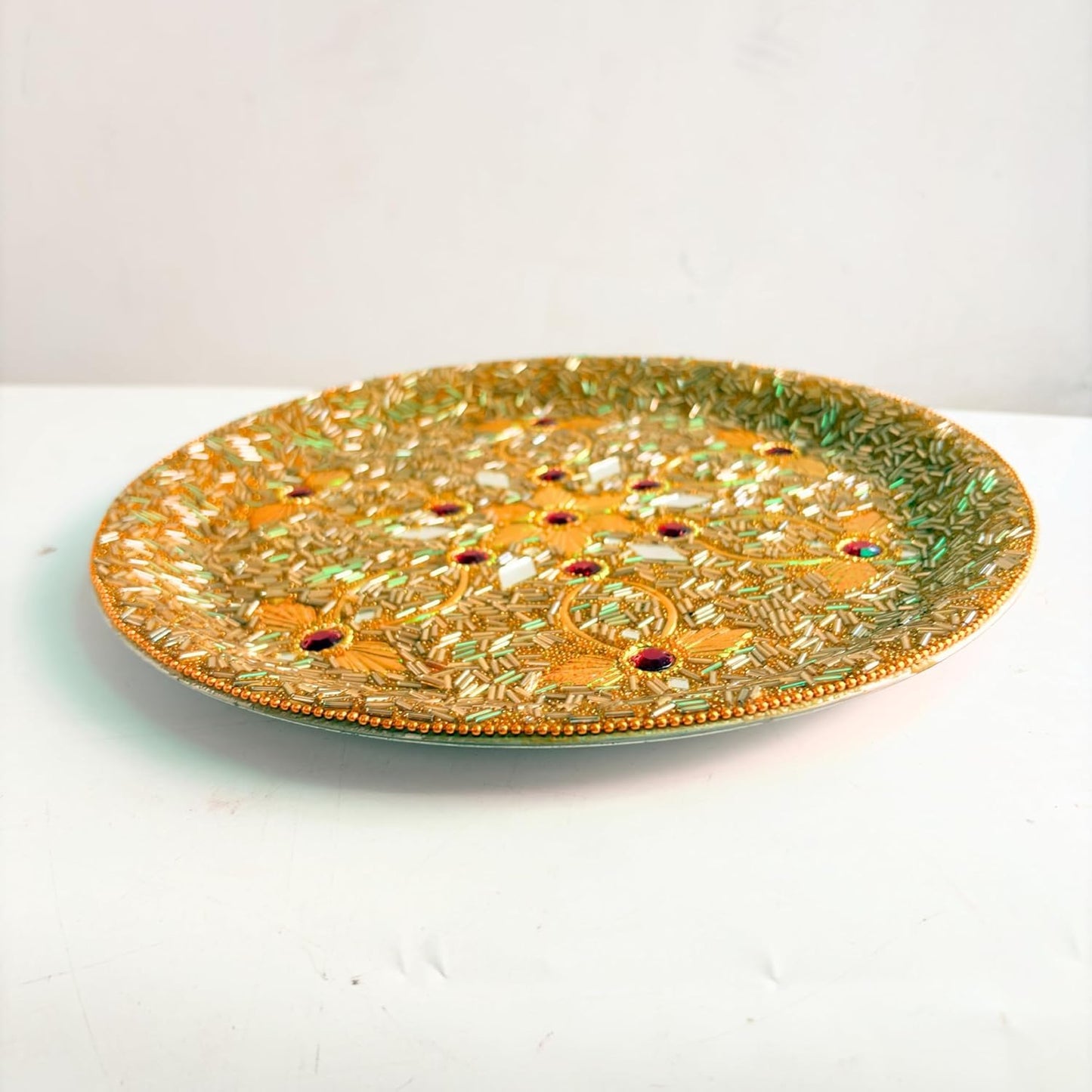 An ornate round decorative tray with intricate gold and green floral patterns and small red jewel accents, displayed on a white surface against a light gray background.