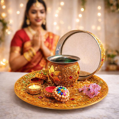 An ornate gold-plated traditional pot with a lid, displayed on a decorated brass tray with a lit candle, red wax, and decorative items. In the softly blurred background, a woman in a red and gold sari is visible with her hands together in a prayer pose.