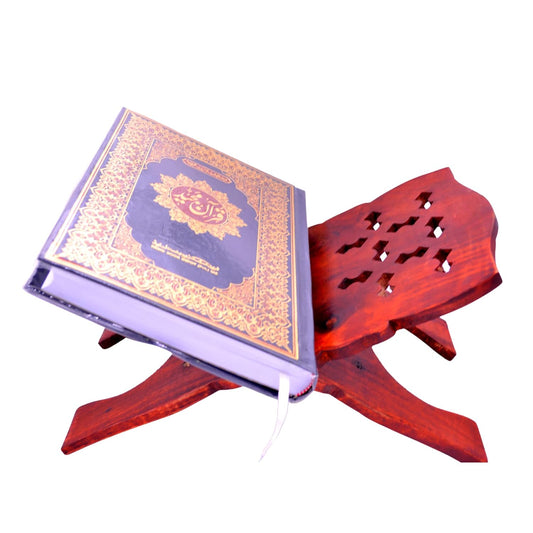A hardcover Quran with ornate gold and blue/purple cover detailing, displayed on a carved reddish-brown wooden stand against a plain white background.