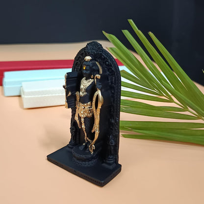 A small black and gold statue of a Hindu deity standing on a decorative arch, displayed on a beige surface with a green palm leaf and stacked boxes in the background.