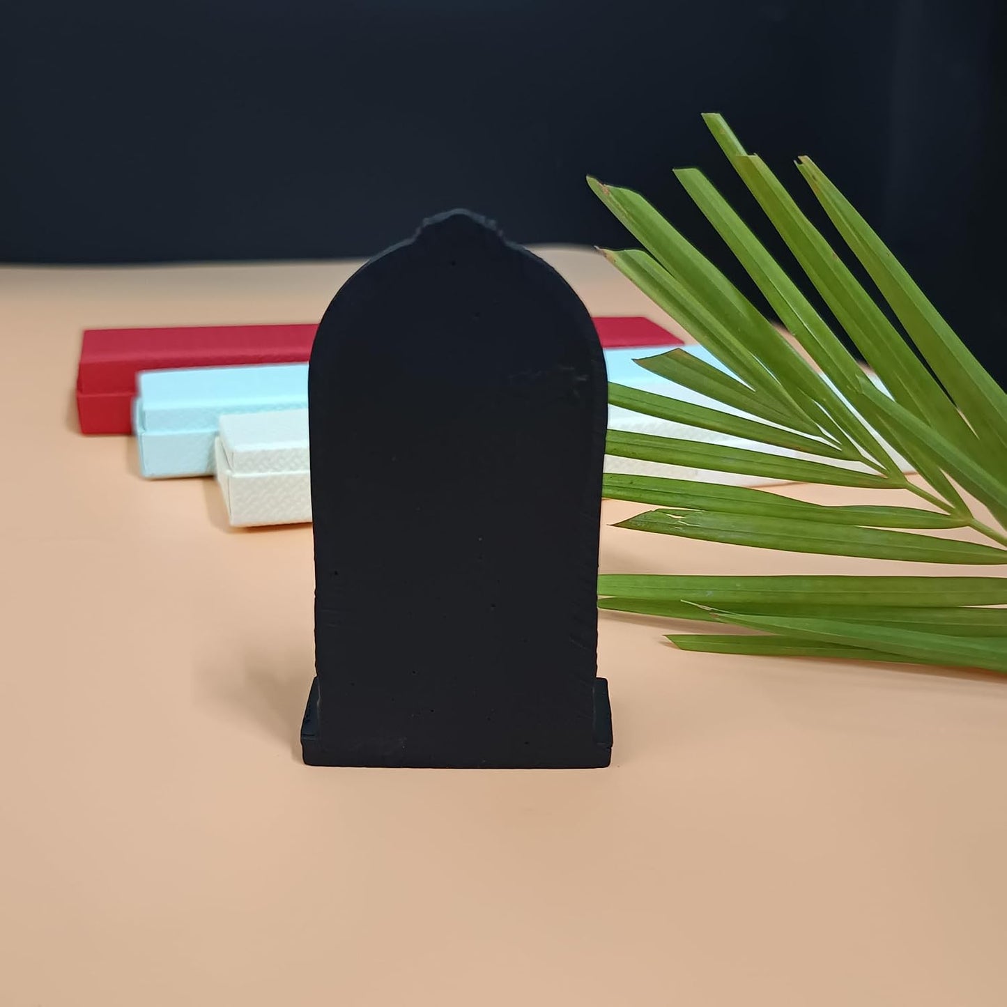 A black tombstone-shaped display stand or plaque holder stands upright on a beige surface, with red, light blue, and white rectangular blocks in the background and green palm leaves on the right.