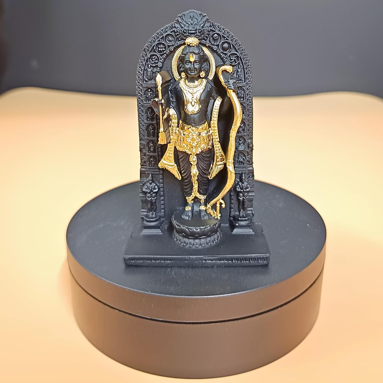 A small black and gold statue of a Hindu deity standing on a round black display base, photographed against a plain beige and dark gray background.