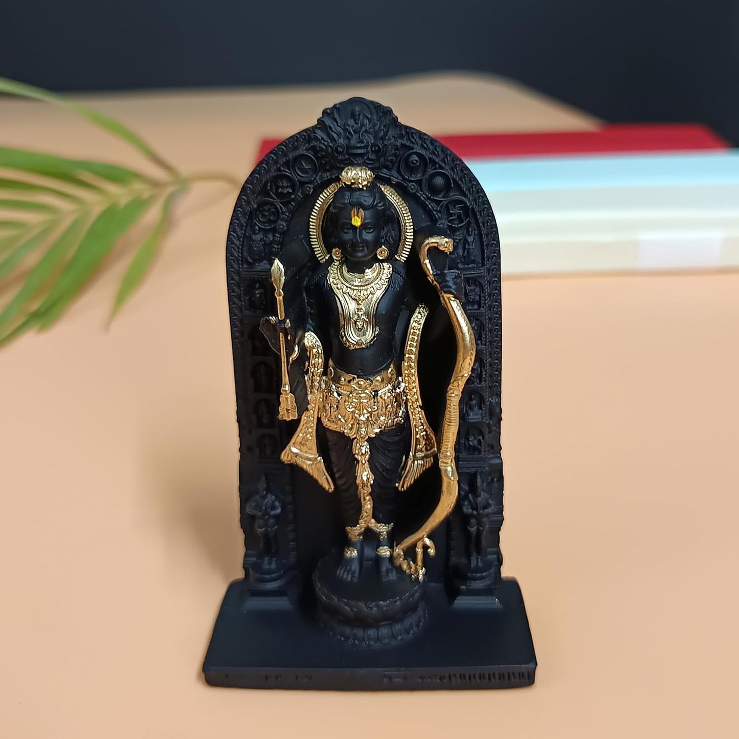 A black and gold Lord Hanuman statue standing in front of an arched black backdrop, displayed on a beige surface with blurred books and a green leaf in the background.
