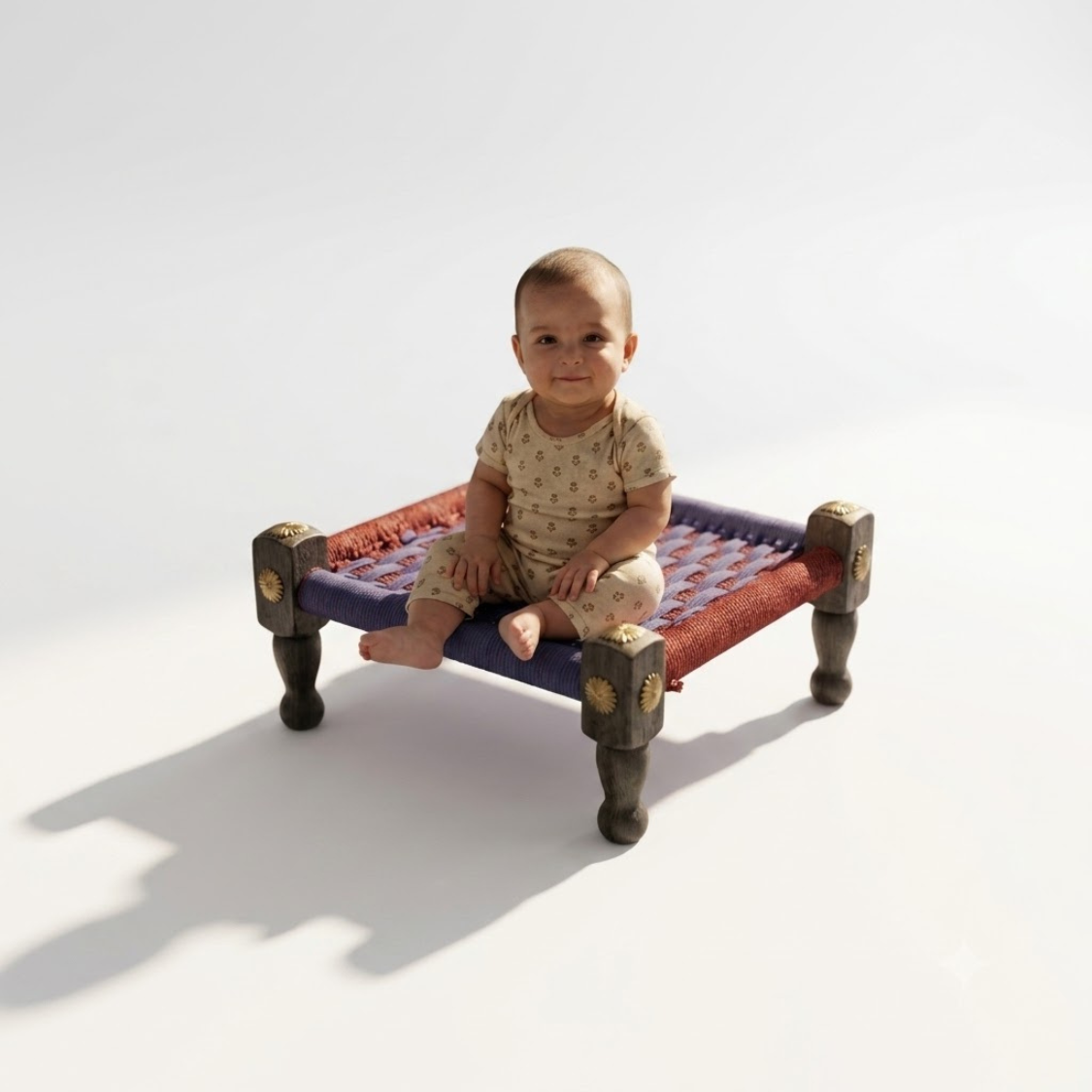 A baby sitting on a traditional Indian wooden charpai bed with a woven red and blue mattress, photographed on a clean white studio background.