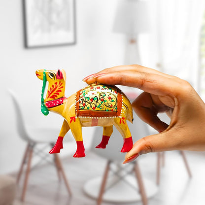 A hand holds a small, brightly painted camel figurine with yellow, red, green, and orange details against a softly blurred indoor background.