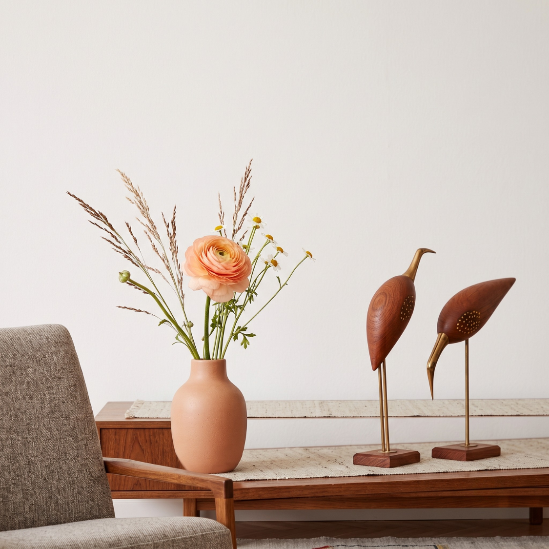 A terracotta-colored ceramic vase filled with dried grasses and a peach ranunculus flower sits on a wooden console table next to two carved wooden bird sculptures on brass stands.