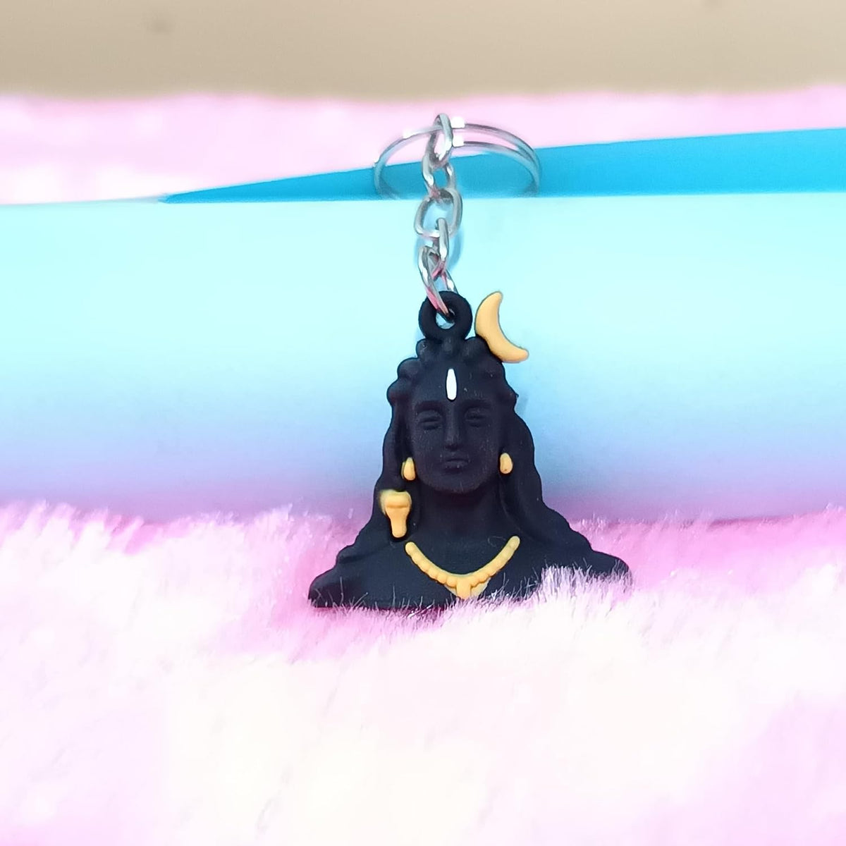 A black Shiva keychain charm with gold and white accents, resting on a pink and blue background.