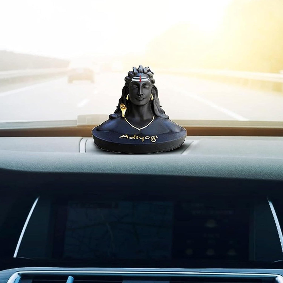 A black resin bust sculpture of Adiyya, a Hindu deity, placed on the dashboard of a car with a blurred highway road and bright sky visible through the windshield.
