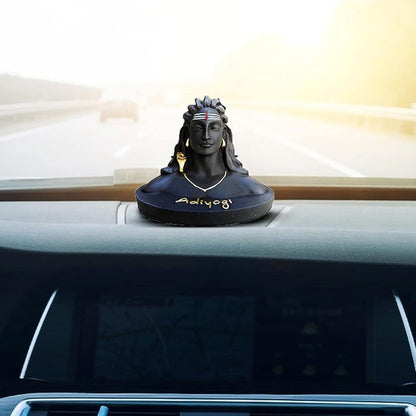 A black resin bust sculpture of Adiyya, a Hindu deity, placed on the dashboard of a car with a blurred highway road and bright sky visible through the windshield.