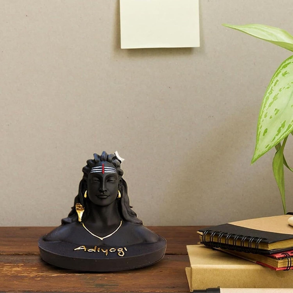 A black resin bust sculpture of Adi Yoga, a Hindu deity, displayed on a wooden table next to books and a green plant against a neutral wall.