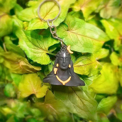 A black Shiva keychain with gold accents, resting on a bed of green leaves.