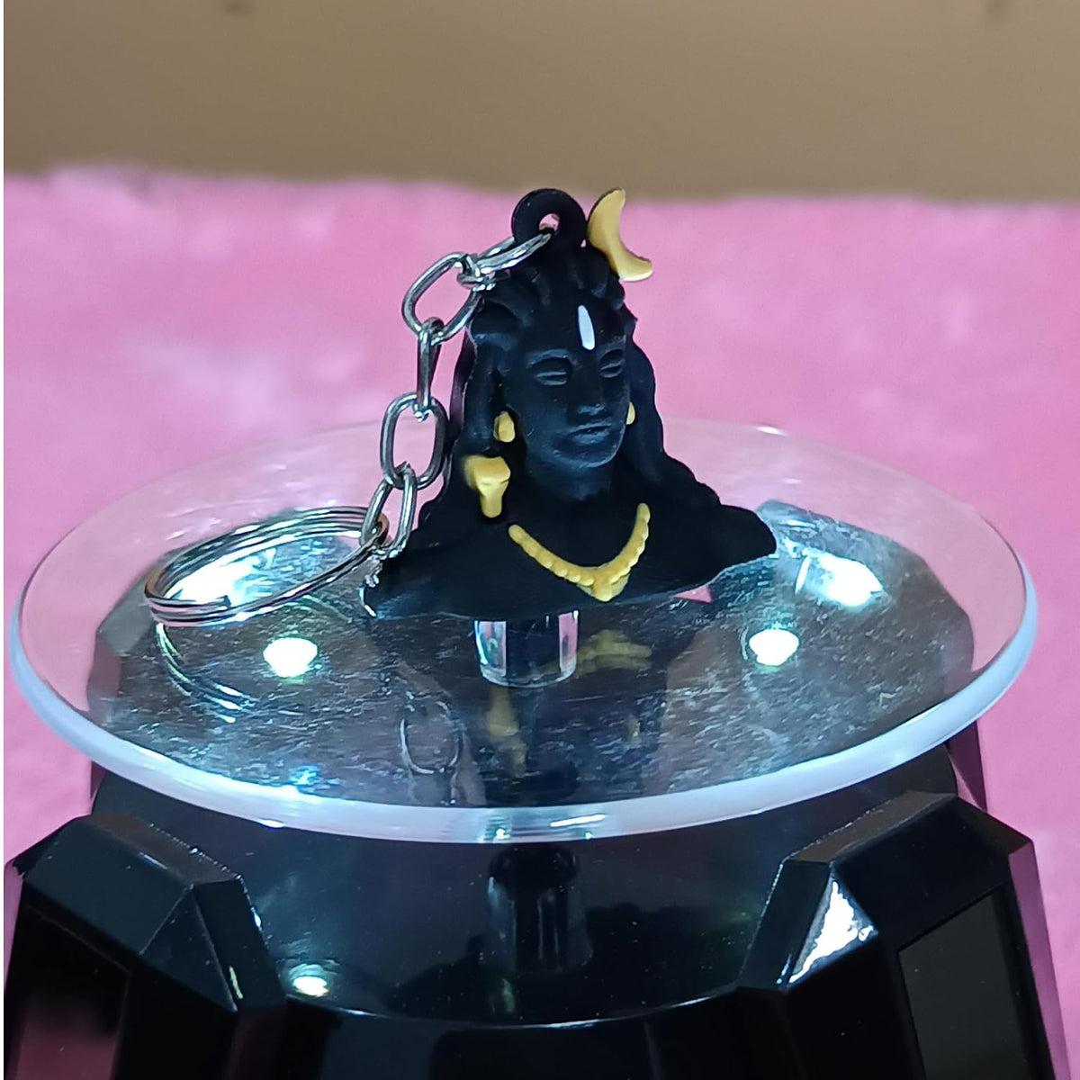 A black Lord Shiva keychain figurine with gold jewelry and a silver keyring, displayed on a clear circular stand with a black base against a pink background.