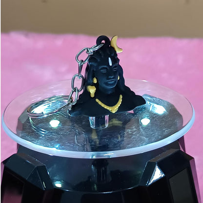 A black Lord Shiva keychain figurine with gold jewelry and a silver keyring, displayed on a clear circular stand with a black base against a pink background.